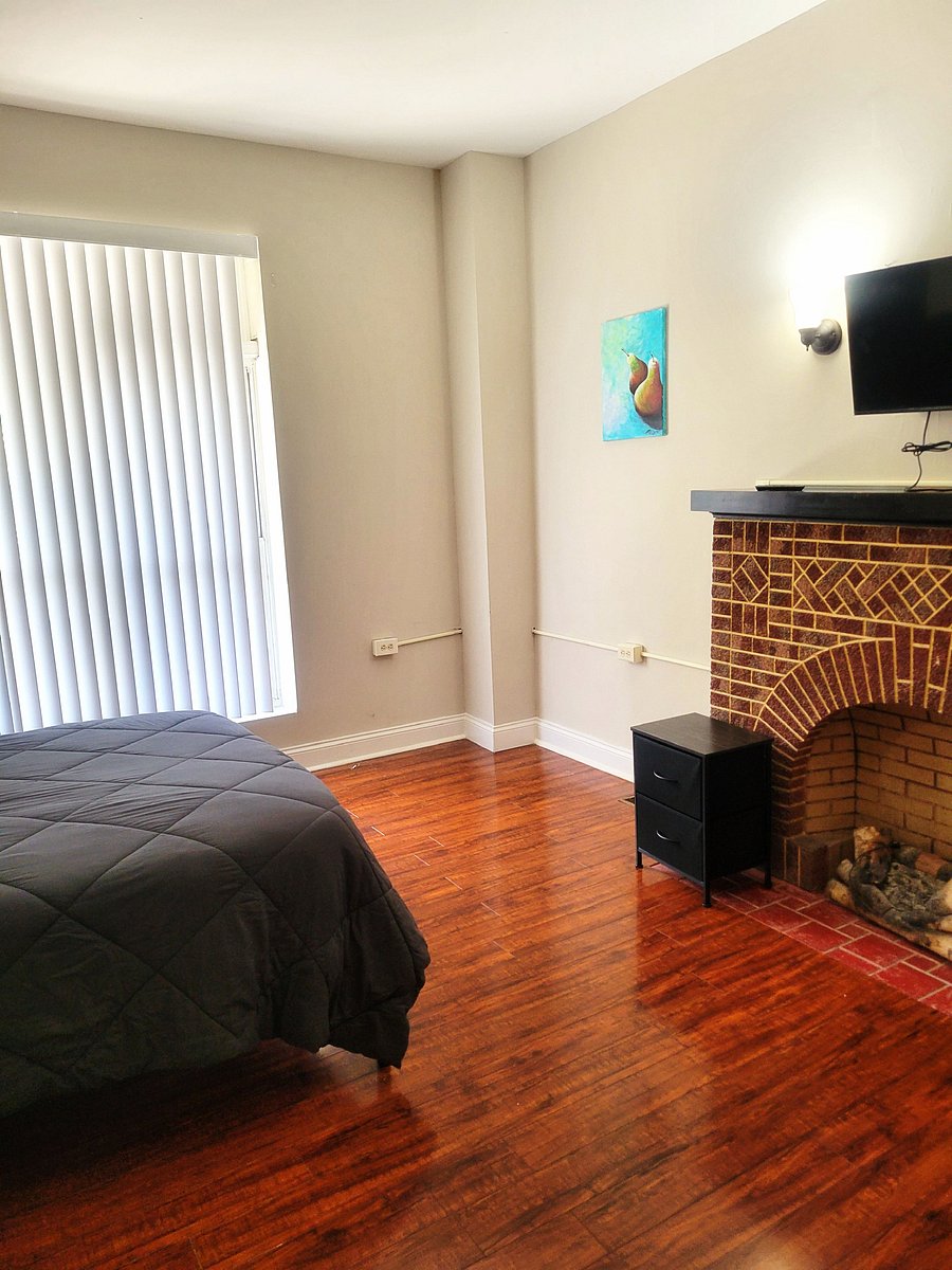 Bedroom with hardwood floors, large window with vertical blinds, and a decorative brick fireplace with mantle and TV mount.