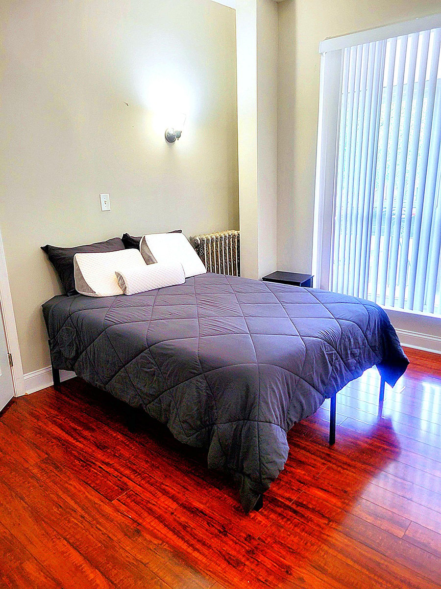 Furnished bedroom with a full bed, hardwood floors, and a large window with vertical blinds for natural light.
