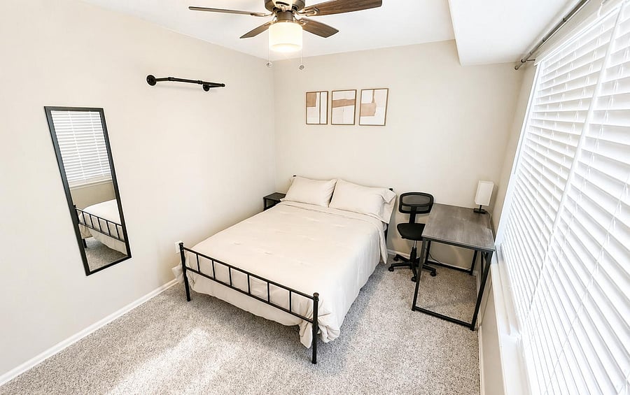 Well-appointed bedroom with a full bed, ceiling fan, large window with blinds, and a dedicated desk workspace.
