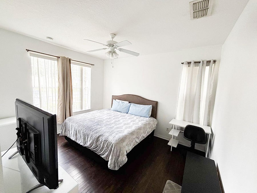 Furnished bedroom with a full bed, ceiling fan, and a dedicated desk area, plus two windows for natural light.