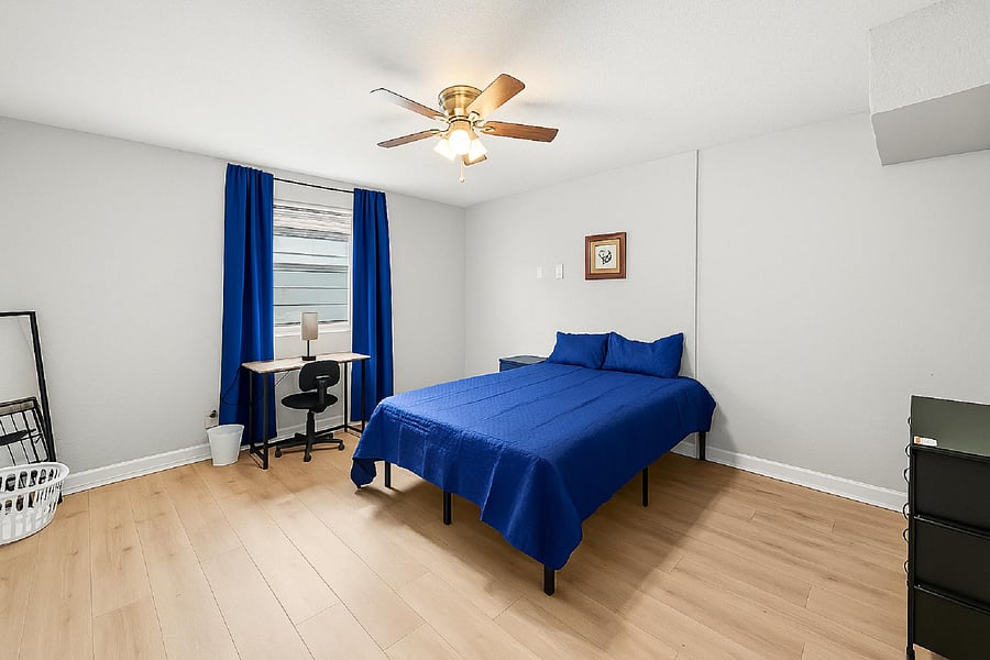 Furnished bedroom with wood-look flooring, ceiling fan, and a dedicated desk area by the window with curtains.