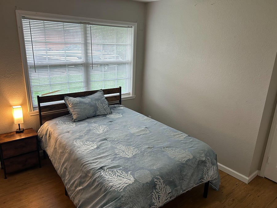 Furnished bedroom with a comfortable bed, nightstand, and large window with blinds for natural light and privacy.