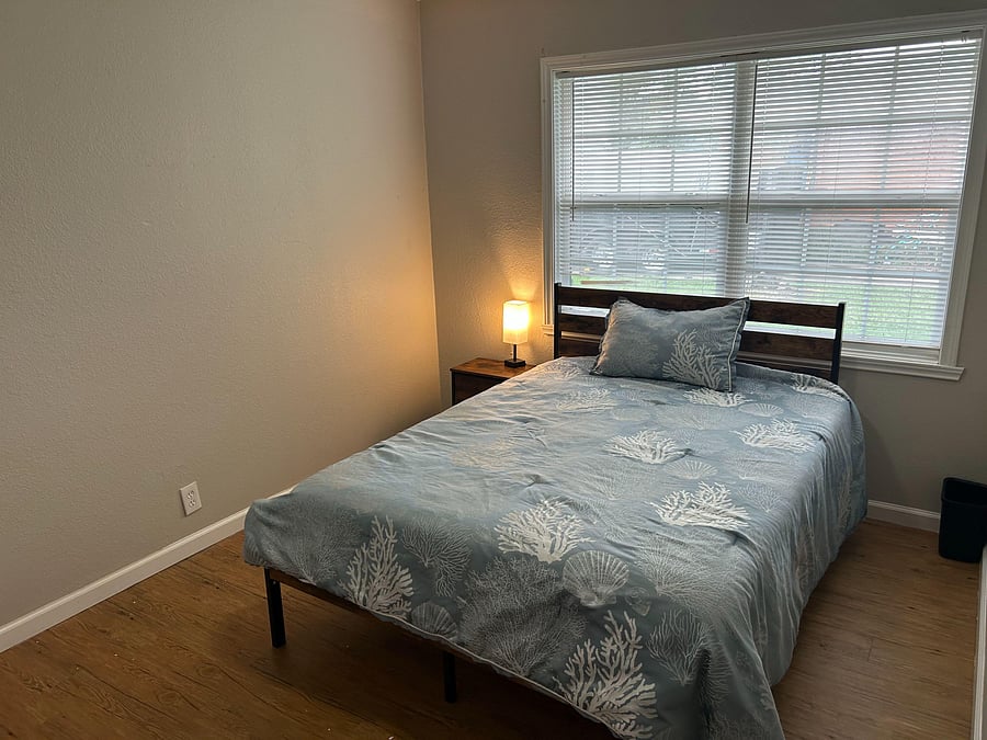 Furnished bedroom with a comfortable bed, nightstand and lamp, plus a large window with blinds for easy light control.