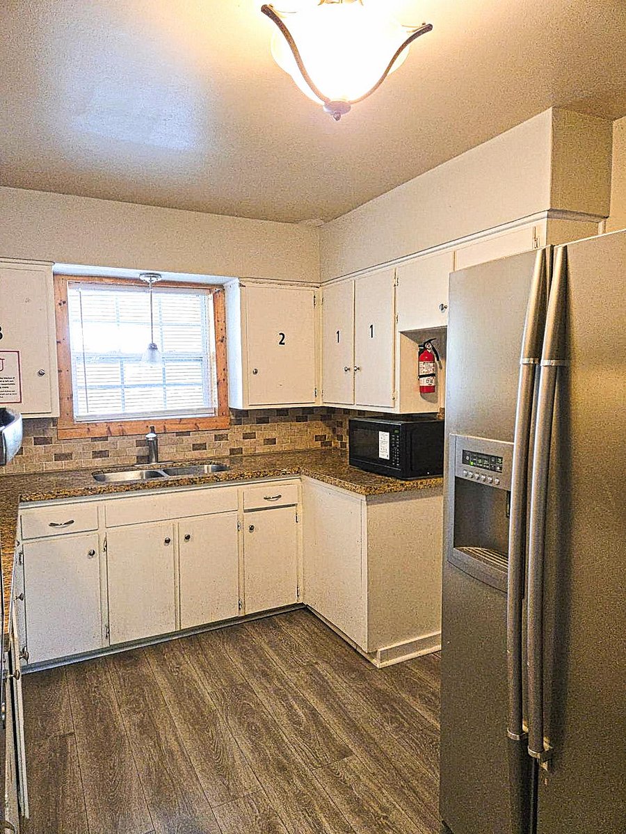 Compact L-shaped kitchen with ample cabinetry, double-basin sink, tile backsplash, and stainless side-by-side fridge.