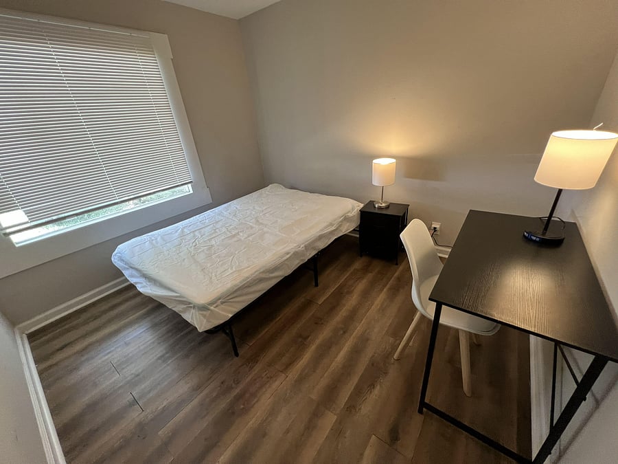 Furnished bedroom with a twin bed, nightstand, and dedicated desk space, plus a large window with blinds.