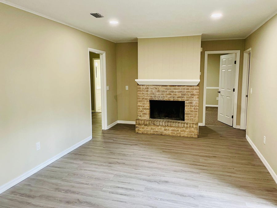 Open living area with durable wood-look flooring and a brick fireplace with mantle, plus convenient access to nearby rooms.