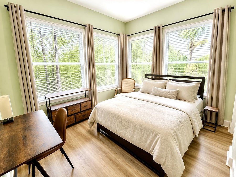 Tranquil bedroom with a full bed, desk/ workspace, and large corner windows with blinds and drapery for natural light.