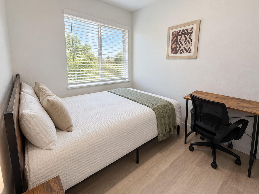 Furnished bedroom with a full bed, large window with blinds, and a dedicated desk workspace for studying or remote work.