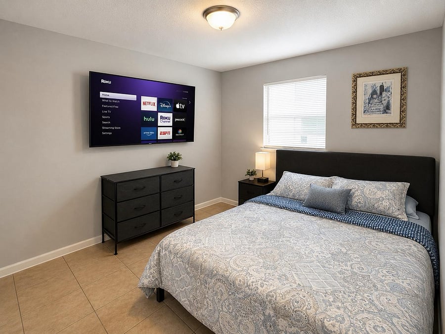 Furnished bedroom with a queen bed, nightstand, dresser, and wall-mounted TV, plus a window with blinds for privacy.