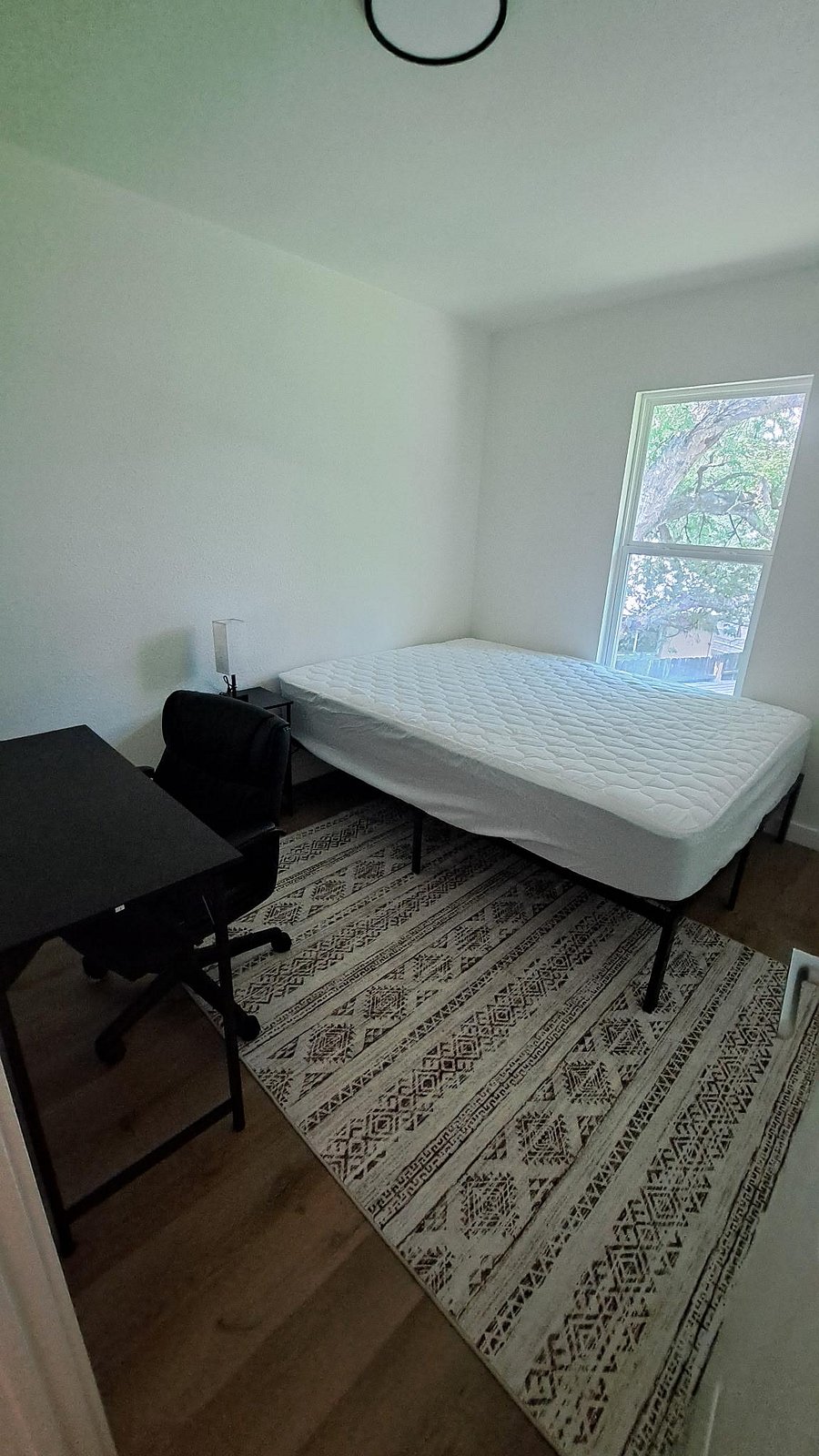 Private bedroom with a large window, twin bed, side table with lamp, and a dedicated desk area for work or study.