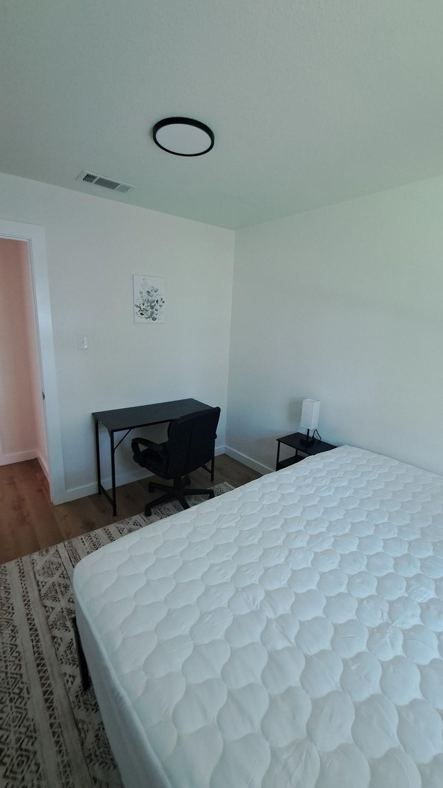 Furnished bedroom with a full bed, nightstand lamp, and a dedicated desk setup, plus durable flooring and clean finishes.