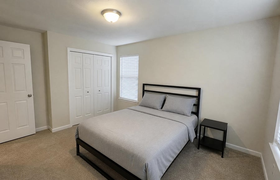 Furnished bedroom with a full bed, nightstand, window with blinds, and a spacious double-door closet.