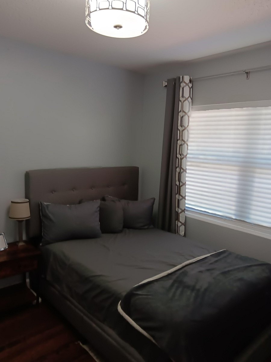 Furnished bedroom with a comfortable bed, upholstered headboard, bedside table, and window with blinds and drapes.