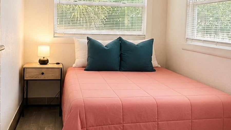 Furnished bedroom with a full-size bed, nightstand lighting, and corner windows with blinds for added privacy.
