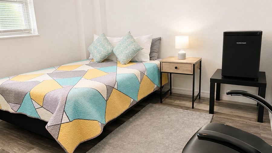 Furnished bedroom with a full bed, nightstand with lamp, mini fridge, and window with blinds for natural light.