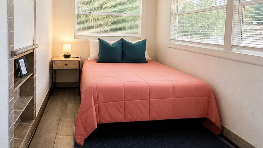 Private bedroom with a full-size bed, nightstand lighting, and three large windows with blinds for natural light.