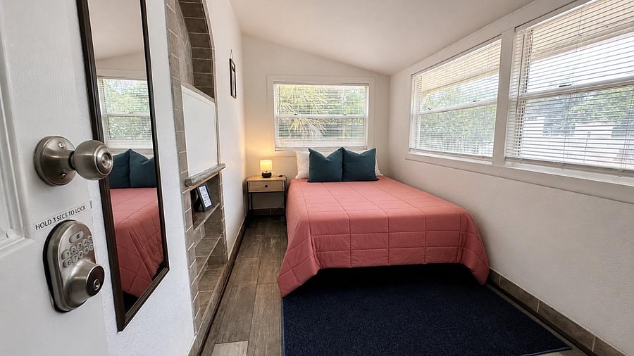 Private bedroom with a full bed, bedside table, and large windows with blinds for natural light and ventilation.