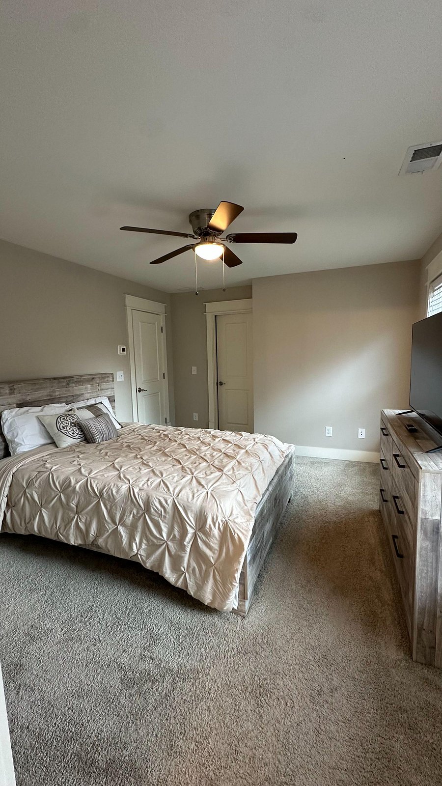 Furnished bedroom with a queen bed, dresser with TV, ceiling fan, and carpeted floors, plus a window with blinds.