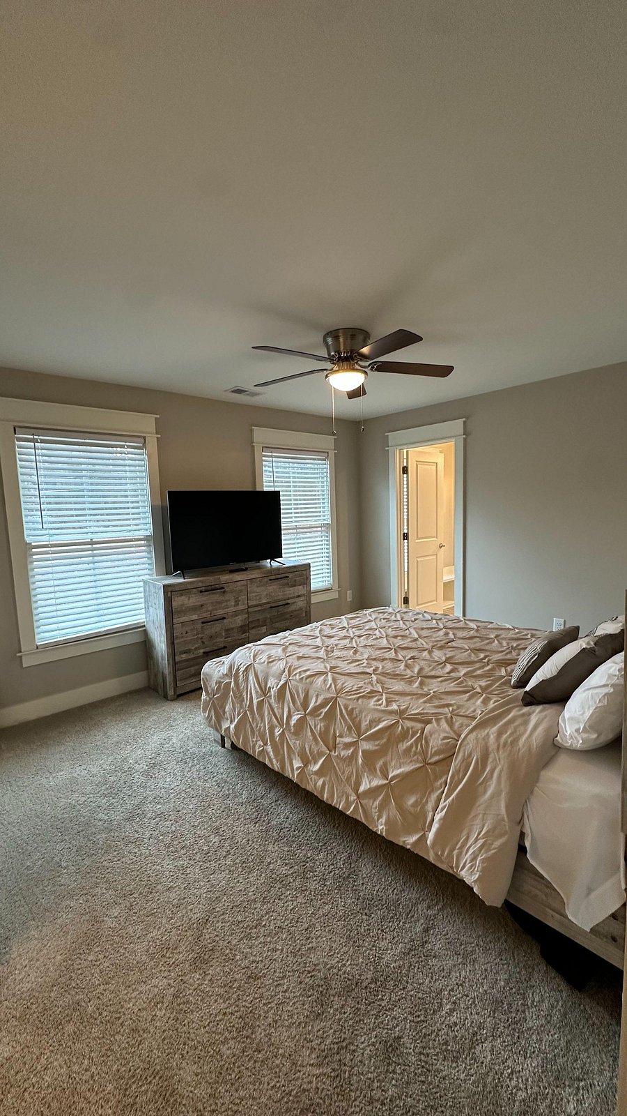 Spacious bedroom with plush carpeting, ceiling fan, and two windows for natural light, plus a dresser and TV setup.
