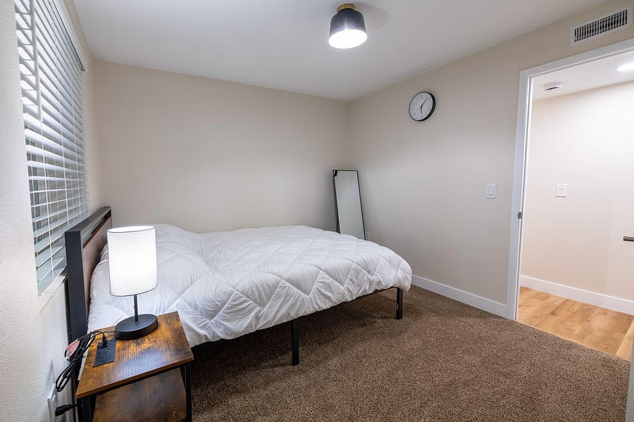 Furnished bedroom with a full bed, nightstand, and window with blinds, plus easy access to the adjacent hallway.