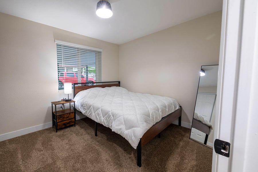Comfortable bedroom with a full bed, nightstand lighting, carpeting, and a large window with blinds for privacy.
