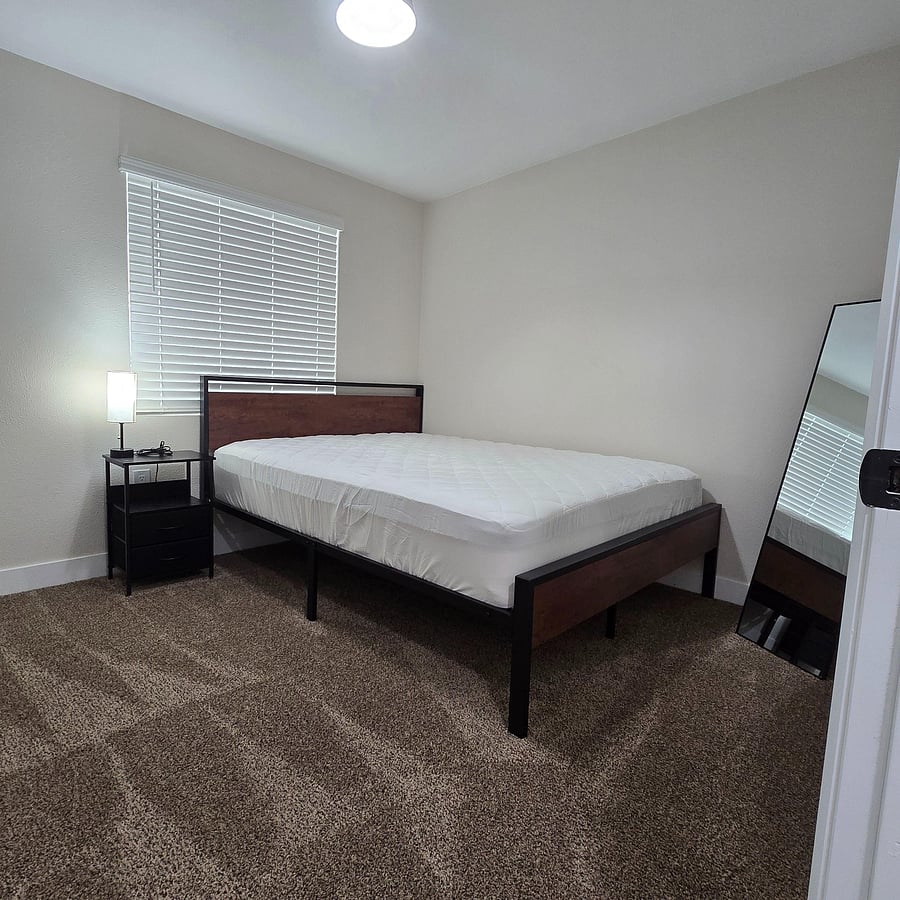 Furnished bedroom with a full-size bed, nightstand and lamp, plus a large window with blinds and carpeted flooring.