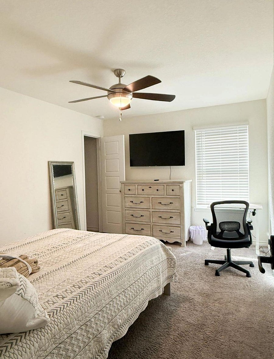 Furnished bedroom with ceiling fan, wall-mounted TV, dresser storage, and a work desk by the window.