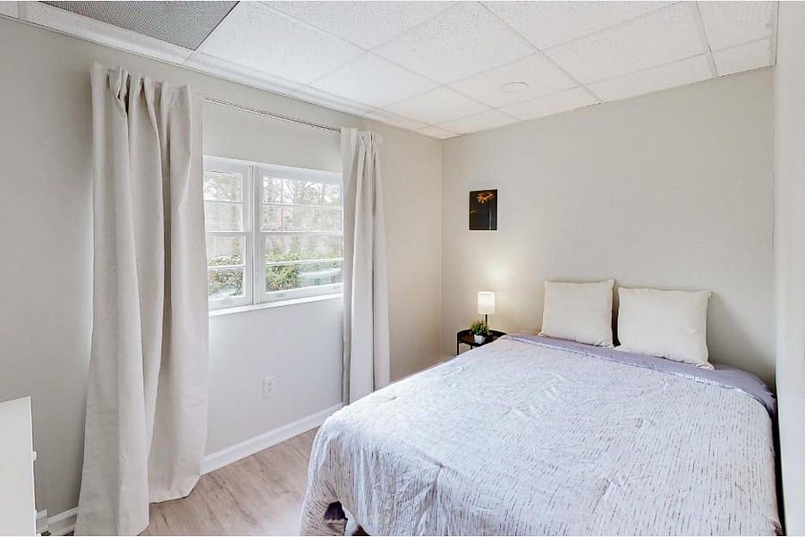 Bedroom offers a comfortable layout with a full-size bed, nightstand lighting, and a large window with curtains.
