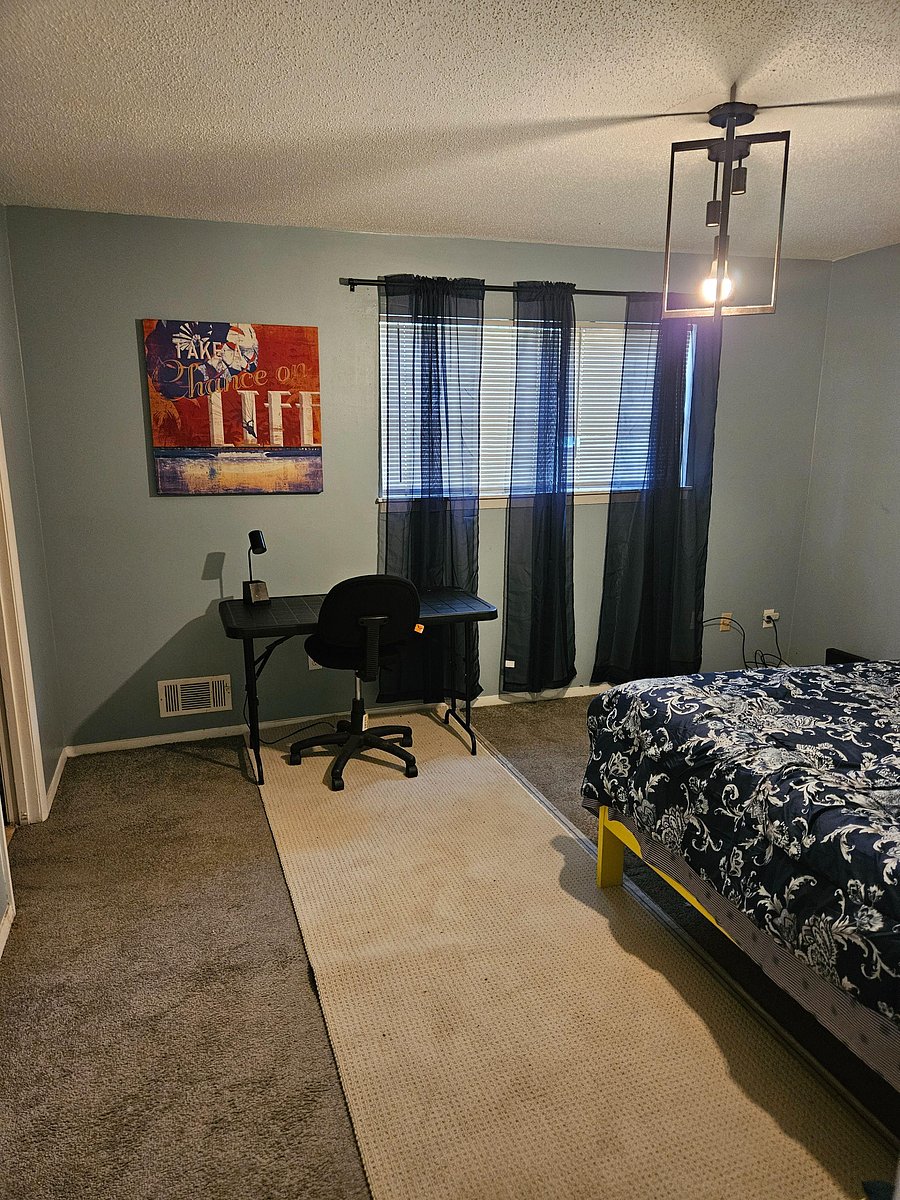 Furnished bedroom with a work desk, large window with blinds, and carpeted flooring for comfortable everyday living.