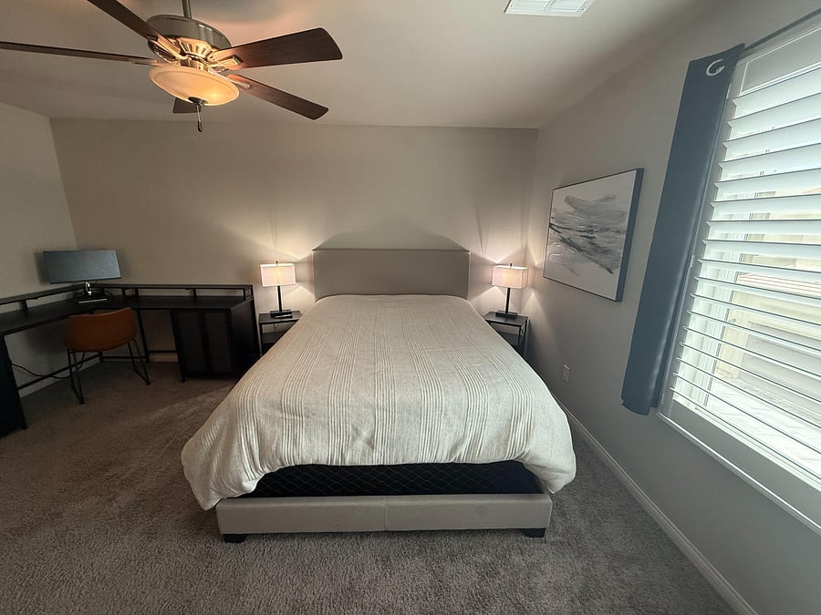 Furnished bedroom with a full bed, bedside lighting, ceiling fan, and a dedicated desk area next to a window with blinds.