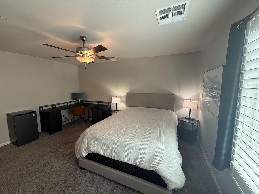 Furnished bedroom with a full bed, nightstands, and a dedicated work area, plus a ceiling fan and window blinds.