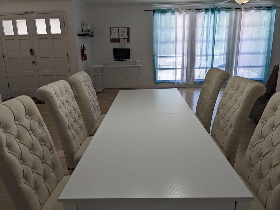 Dining area features a long table with upholstered seating and large windows with curtains for comfortable shared meals.