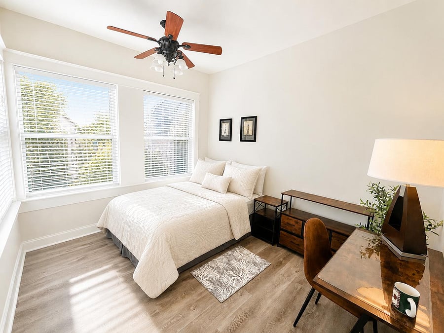 Sunlit bedroom with large windows, ceiling fan, and wood-look flooring, plus a desk area and storage for easy living.