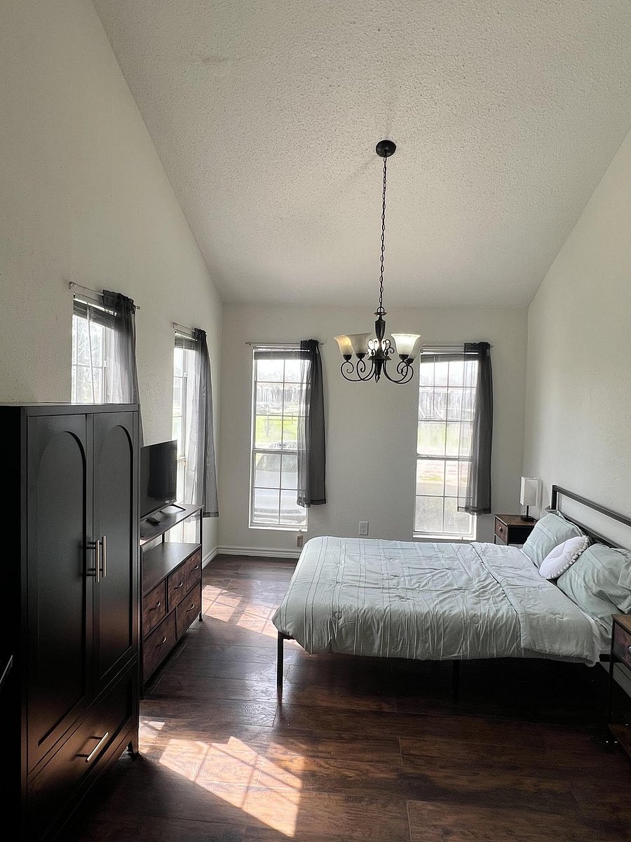 Spacious bedroom with dark wood floors, three large windows, a bed, TV setup, and ample storage with dresser and armoire.