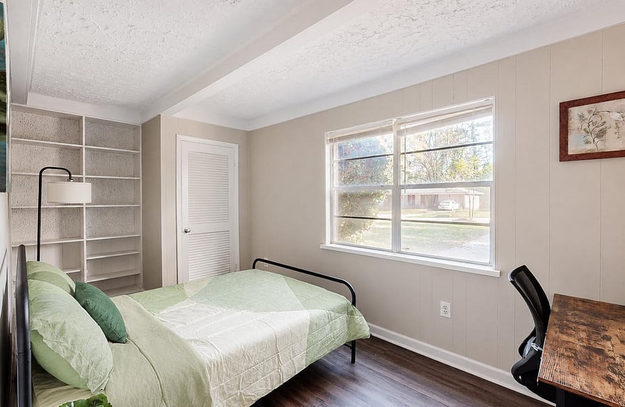Furnished bedroom with a full bed, large window for natural light, built-in shelving, and a dedicated desk workspace.