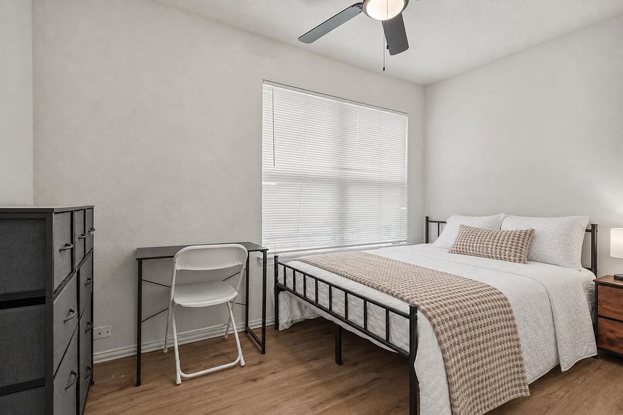 Furnished bedroom with a full bed, dresser, and compact desk setup, plus a large window with blinds and ceiling fan.