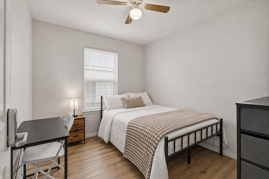 Furnished bedroom with a full bed, desk workspace, dresser storage, and a window with blinds plus ceiling fan light.