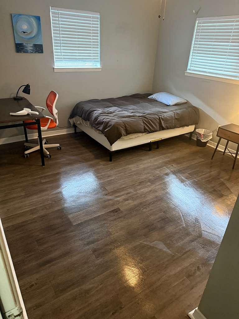 Private bedroom with wood-look flooring, two windows with blinds, and space for a bed plus a work desk.