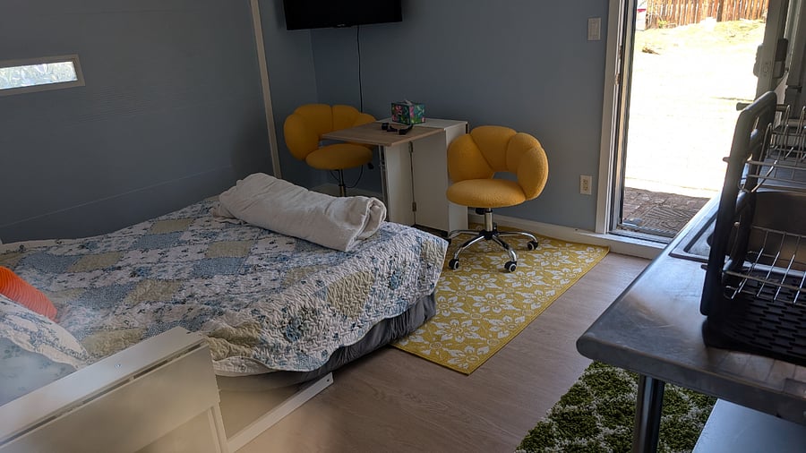 This gives you a bit of a view. Your front door is open to let. In some light. You can see a little bit of the kitchen off to the right side. You can see the bed is a large part of the space. And then the small chairs and table.
