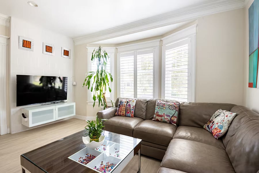 Living Room: Unwind in a sun-filled living space featuring a  genuine leather sectional and a sleek 55 wall-mounted smart TV set on a refined accent wall. Rich Craftsman-inspired flooring adds depth and warmth, while three signature Craftsman-style windows bring in abundant natural light, creating a cozy yet open ambiance