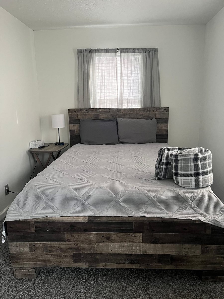 Spacious master bedroom with natural light, featuring a sturdy rustic bed, bedside table, lamp, fan, and 32-inch smart TV.