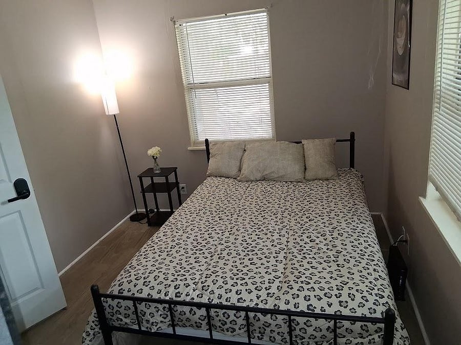 Private bedroom features a full-size bed, side table, and two windows with blinds for natural light and privacy.