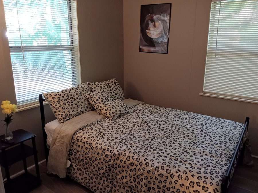 Private bedroom features a full-size bed, side table, and two windows with blinds for natural light and privacy.