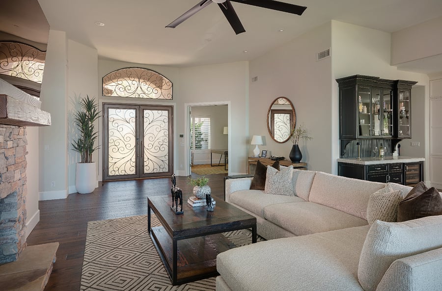 The main living area features a floor-to-ceiling stacked stone fireplace, large format flat screen TV, custom built-in shelving, and a oversized sectional with chaise. Soaring ceilings, dark hardwood floors, and sliding doors opening to the patio complete the space.