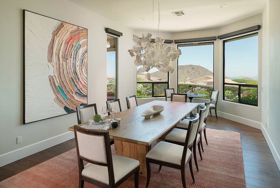 The dining room features two dedicated seating areas: a formal eight-seat solid wood table with upholstered nailhead chairs, sculptural chandelier, and Troon Mountain views, plus a bistro table for two tucked into the bay window with direct views of city lights and desert sunsets.