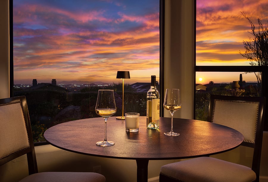 The dining room features two dedicated seating areas: a formal eight-seat solid wood table with upholstered nailhead chairs, sculptural chandelier, and Troon Mountain views, plus a bistro table for two tucked into the bay window with direct views of city lights and desert sunsets.