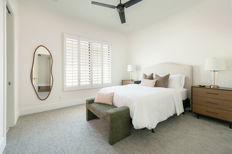 Queen bedroom with upholstered headboard, bench, dual nightstands, plantation shutters, ceiling fan, and sliding door closet.