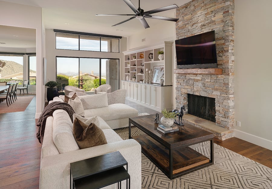 The main living area features a floor-to-ceiling stacked stone fireplace, large format flat screen TV, custom built-in shelving, and a oversized sectional with chaise. Soaring ceilings, dark hardwood floors, and sliding doors opening to the patio complete the space.