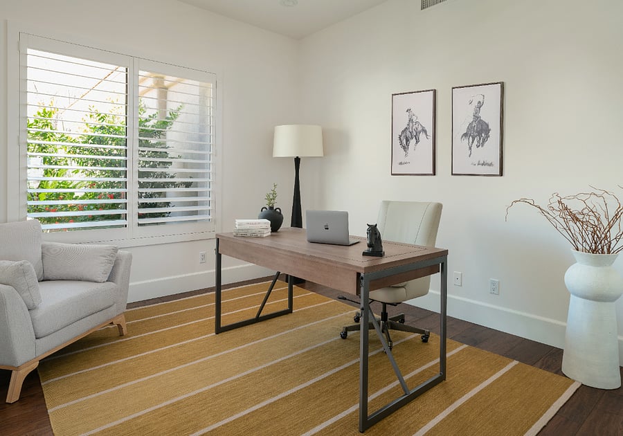Dedicated home office with a full-size desk, adjustable office chair, plantation shutters, and a separate seating area. A private, well-lit workspace with natural light and garden views.
