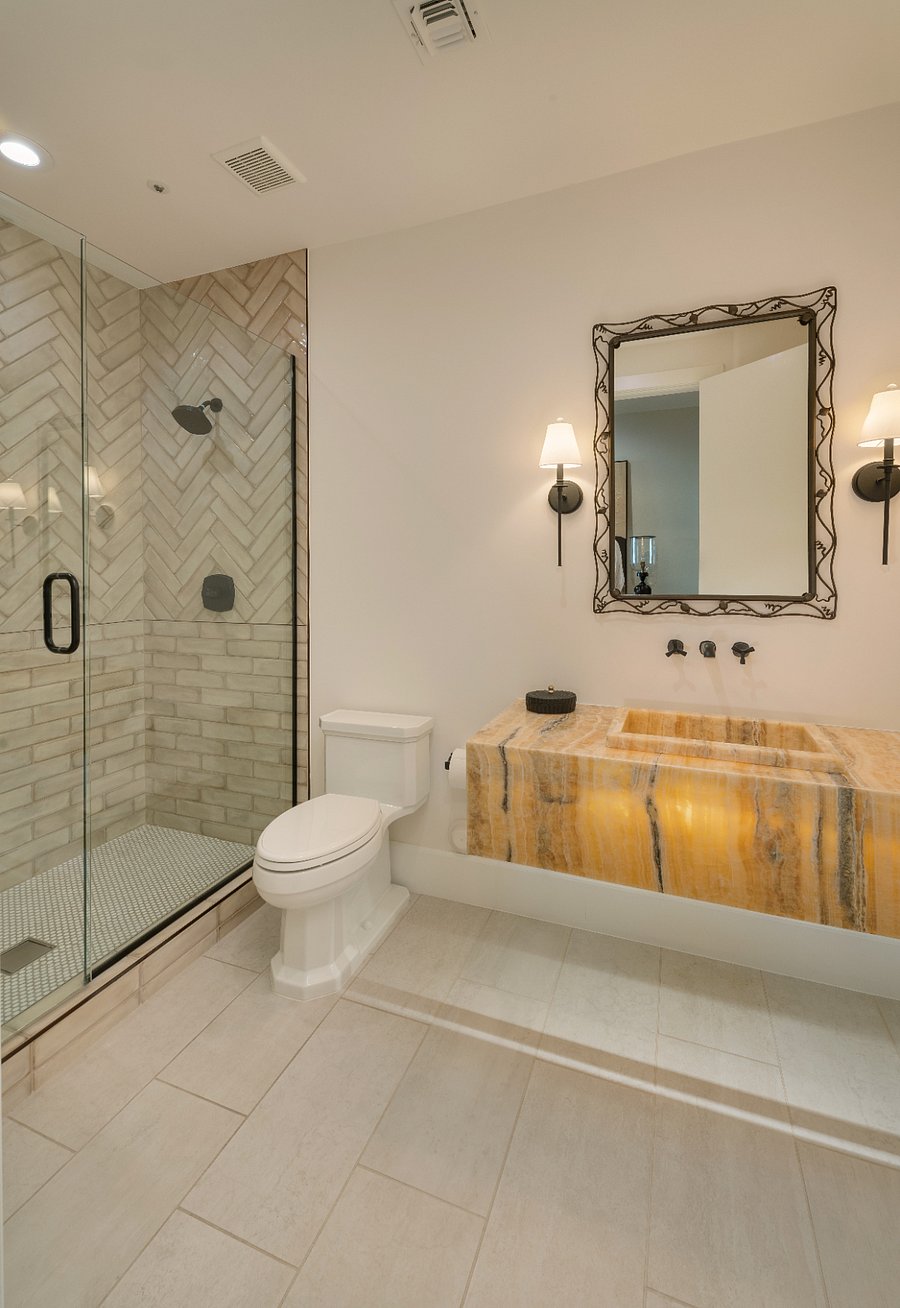 Full hallway bathroom with a glass-enclosed shower with herringbone tile, ornate framed mirror, wall-mounted matte black fixtures, and a carved onyx stone vanity sink.